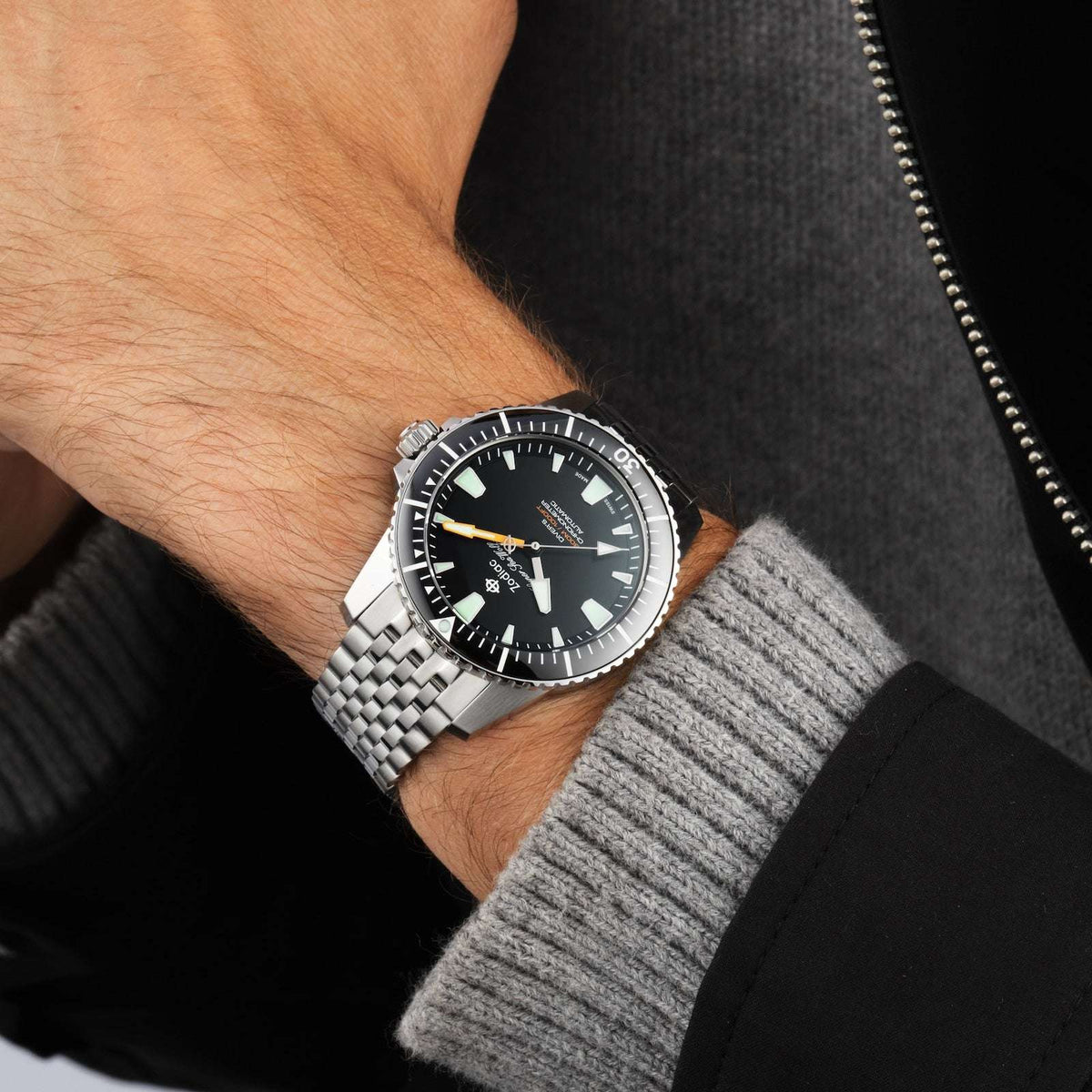 Orologio subacqueo in acciaio al polso, quadrante nero con indici verdi e lancette arancioni.