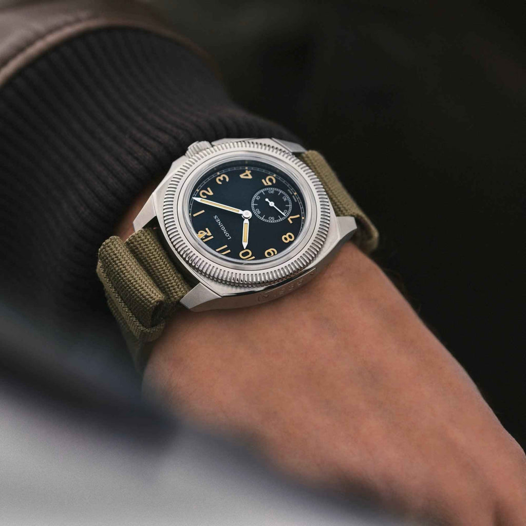 Orologio Longines su polso con quadrante nero e cinturino marrone in tessuto.