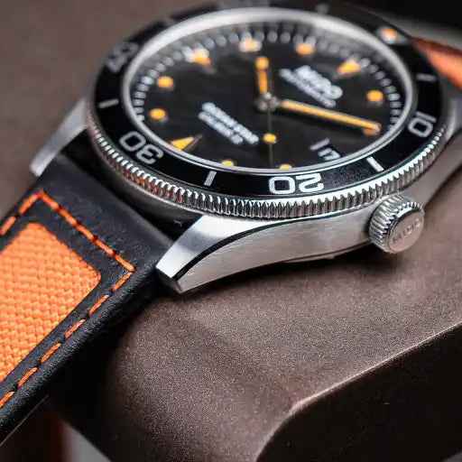 Orologio con ghiera nera e corona laterale; cinturino nero con cuciture arancioni.